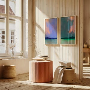 K&ouml;nnte beinhalten: Zwei gerahmte abstrakte Landschaftsbilder mit blauen, gr&uuml;nen und orangen Farbt&ouml;nen h&auml;ngen an einer hellen Wand mit Holzvert&auml;felung. Ein korallenfarbener, gerippter zylindrischer Hocker und ein Weidenkorb stehen auf einem Holzboden. Ein Fenster ist sichtbar.