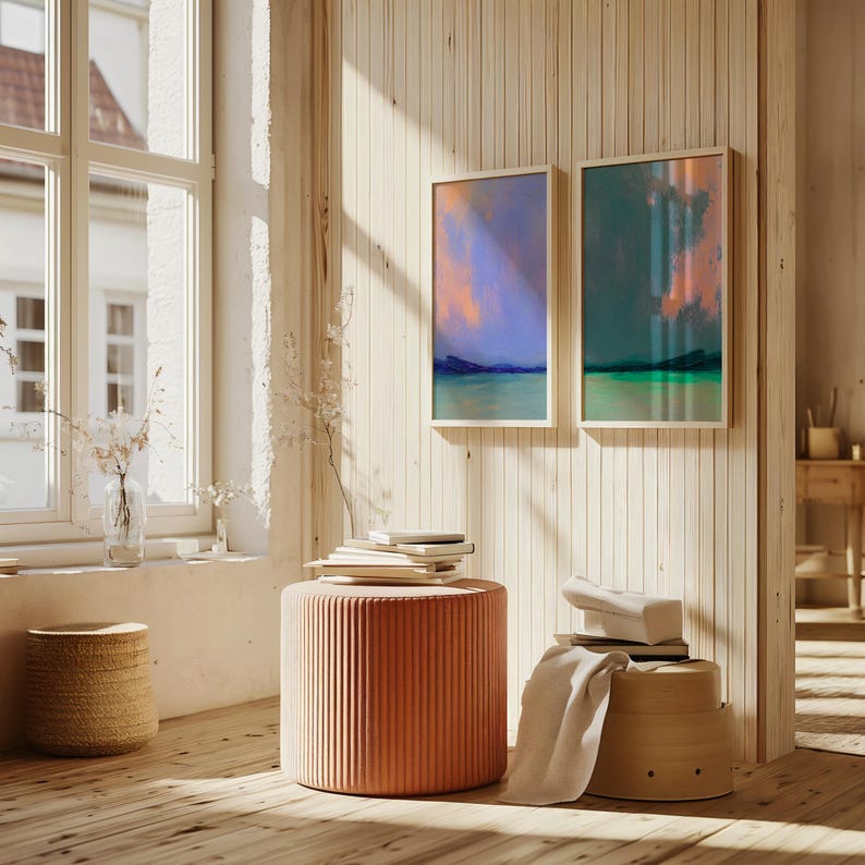 K&ouml;nnte beinhalten: Zwei gerahmte abstrakte Landschaftsbilder mit blauen, gr&uuml;nen und orangen Farbt&ouml;nen h&auml;ngen an einer hellen Wand mit Holzvert&auml;felung. Ein korallenfarbener, gerippter zylindrischer Hocker und ein Weidenkorb stehen auf einem Holzboden. Ein Fenster ist sichtbar.