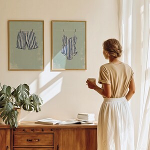 May include: Two framed prints depict blue and white striped swimwear hanging on a clothesline against a light green background. The prints are in gold frames. A woman stands nearby, holding a cup, with a wooden cabinet and a potted plant in the foreground.