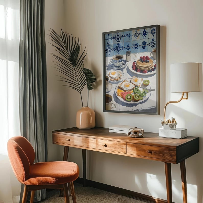 Puede incluir: Una impresi&oacute;n art&iacute;stica enmarcada representa una escena de desayuno con panqueques, huevos, fruta y caf&eacute;. La obra de arte cuelga sobre un escritorio de madera con una silla color melocot&oacute;n. Tambi&eacute;n se ve una l&aacute;mpara con una pantalla blanca y un jarr&oacute;n con una hoja de palma.