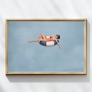 May include: A framed wall art print featuring a figure in a coral swimsuit and black sunglasses lounging on a blue and white striped inflatable pool ring, set against a solid light blue background with subtle circular ripples in the water.