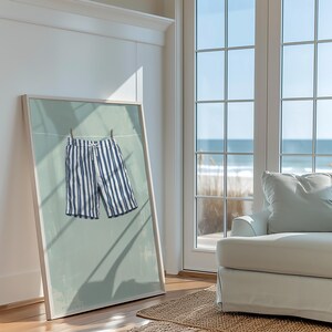 May include: Framed art print featuring a pair of blue and white striped shorts hanging on a clothesline. The artwork has a light blue background and is displayed in a room with a view of the ocean.