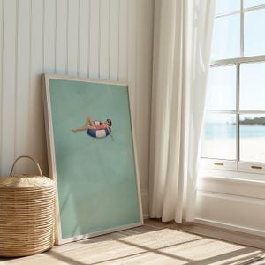 Pu&ograve; includere: Una stampa artistica incorniciata che presenta un'illustrazione minimalista di una persona con costume da bagno rosa e occhiali da sole, sdraiata su un salvagente gonfiabile a righe blu e bianche al centro di uno sfondo verde salvia tenue.