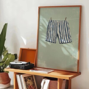 May include: A framed print featuring blue and white striped swim trunks hanging on a clothesline against a muted green background. The artwork is displayed on a wooden table with a record player and a plant.