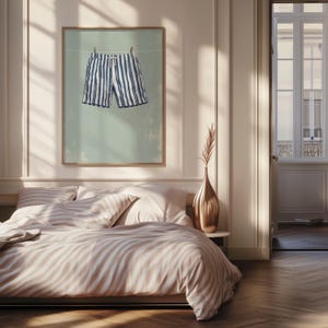 May include: A framed print featuring blue and white striped shorts hanging on a clothesline against a muted green background. The artwork is displayed above a bed with striped bedding, creating a cohesive and stylish bedroom aesthetic.