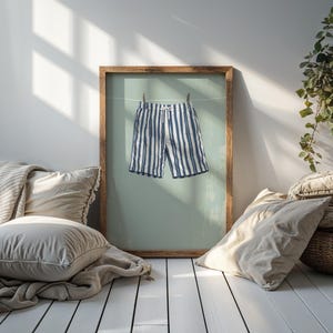 May include: A framed print featuring a pair of blue and white striped shorts hanging on a clothesline. The shorts are the focal point, displayed against a light green background within a wooden frame. The scene is set in a bright, airy room.