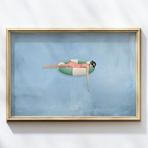 Pode incluir: Uma impressão artística emoldurada retrata uma pessoa flutuando em uma piscina em um anel inflável listrado verde e branco. A pessoa está usando óculos de sol e um maiô rosa. O fundo é uma superfície azul clara texturizada.