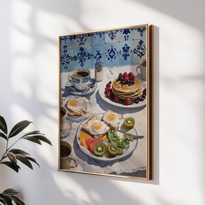 Puede incluir: Pintura enmarcada de una escena de desayuno. La pintura representa una mesa con panqueques, huevos, fruta y caf&eacute;. El fondo presenta azulejos azules y blancos estampados. La obra de arte est&aacute; enmarcada en un marco dorado.