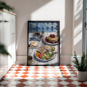 Puede incluir: Una impresi&oacute;n enmarcada de una escena de desayuno con panqueques con bayas, huevos fritos, tostadas, fruta y caf&eacute;. La obra de arte tiene un mantel blanco, azulejos azules y blancos y un marco negro. El suelo es a cuadros rojos y blancos.