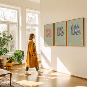 May include: Three framed art prints of shorts hanging on a clothesline. The shorts are red and white striped, navy and white striped, and blue and white striped. The prints are in a room with a woman wearing a mustard-colored coat.