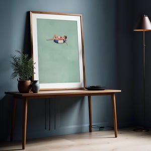 Pu&ograve; includere: Una stampa artistica incorniciata che mostra un'illustrazione minimalista di una figura in costume da bagno color corallo e occhiali da sole che si rilassa su un salvagente a righe blu e bianche, su sfondo verde salvia in una cornice in legno chiaro.