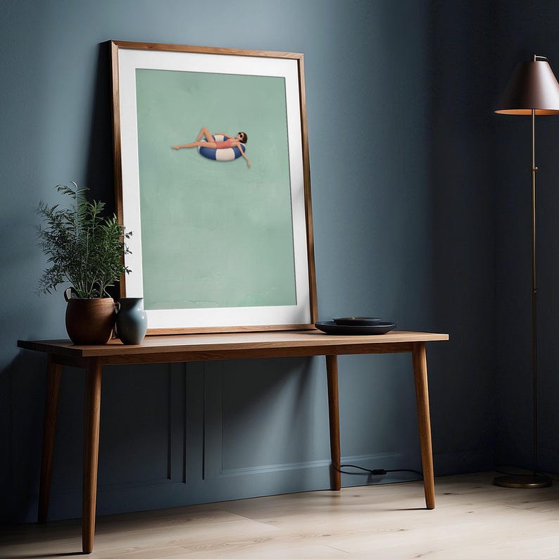 Pu&ograve; includere: Una stampa artistica incorniciata che mostra un'illustrazione minimalista di una figura in costume da bagno color corallo e occhiali da sole che si rilassa su un salvagente a righe blu e bianche, su sfondo verde salvia in una cornice in legno chiaro.