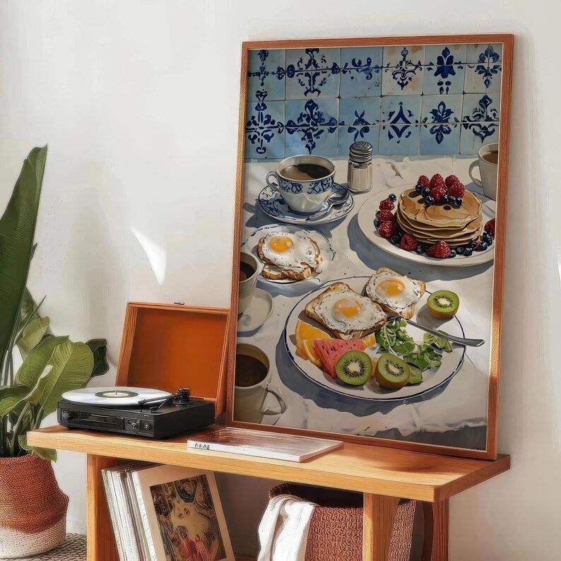 Puede incluir: Una impresi&oacute;n enmarcada de una escena de desayuno con panqueques, huevos, fruta y caf&eacute;. La obra de arte se exhibe en una estanter&iacute;a de madera con un tocadiscos y libros. El fondo presenta azulejos estampados azules y blancos.