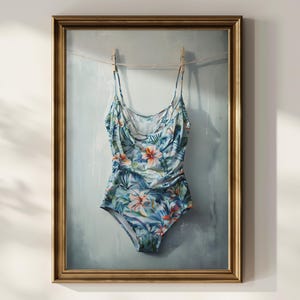 May include: A framed artwork featuring a one-piece swimsuit with a tropical floral pattern. The swimsuit, in shades of blue, green, and orange, is suspended from a clothesline with wooden clothespins against a light blue backdrop.