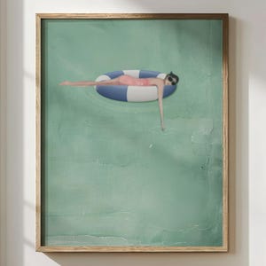 May include: A framed art print depicts a person floating on a blue and white striped inflatable ring in a pool. The person wears a pink swimsuit and sunglasses. The background is a textured, light green color.