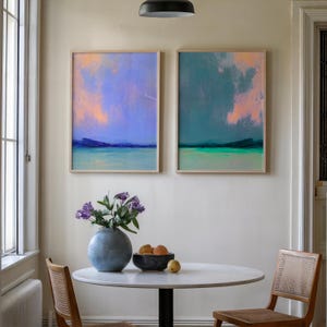 K&ouml;nnte beinhalten: Zwei gerahmte abstrakte Landschaftsbilder in Blau-, T&uuml;rkis- und Pfirsicht&ouml;nen h&auml;ngen an einer cremefarbenen Wand. Ein runder Tisch mit Marmorplatte, eine blaue Vase mit lila Blumen und eine Schale mit Obst vervollst&auml;ndigen die Szene. Holzst&uuml;hle mit gewebten R&uuml;cken umrunden den Tisch.
