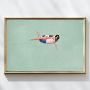 May include: A framed horizontal wall art print featuring a stylized illustration of a person with dark hair and sunglasses wearing a pink swimsuit, floating on a blue and white striped circular pool tube in calm, light green water.