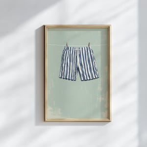 May include: Framed art print featuring blue and white striped swim trunks hanging on a clothesline. The shorts are suspended by wooden clothespins against a muted green background. The artwork is encased in a gold-colored frame.