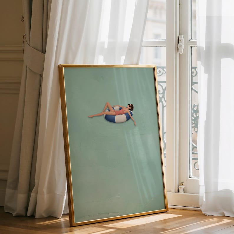 Pu&ograve; includere: Una stampa artistica incorniciata con sfondo verde salvia che presenta una piccola illustrazione di una persona in costume da bagno rosa e occhiali da sole, sdraiata su un salvagente circolare a righe blu e bianche al centro.