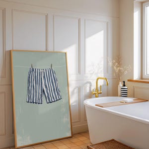 May include: A framed print featuring blue and white striped swim trunks hanging on a clothesline. The artwork has a light green background and a wooden frame. The scene evokes a relaxed, summery vibe, suitable for bathroom or beach-themed decor.
