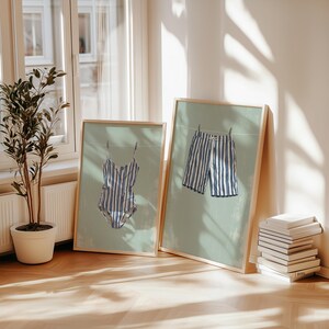 May include: Two framed art prints featuring blue and white striped swimwear. One print shows a swimsuit, the other shows shorts, both hanging on a clothesline against a light green background. The frames are light wood.