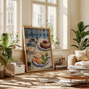 Puede incluir: Una impresi&oacute;n enmarcada de una escena de desayuno con panqueques con bayas, huevos fritos, fruta y caf&eacute;. La obra de arte tiene un marco de madera clara y est&aacute; apoyada contra una pared en una habitaci&oacute;n soleada con un sof&aacute; y plantas.
