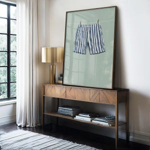 May include: A framed art print featuring blue and white striped shorts hanging on a clothesline against a muted green background. The artwork is displayed on a wooden console table with a gold lamp.