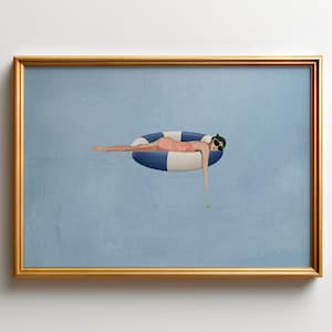 May include: A framed wall art print featuring a figure in a pink swimsuit and black sunglasses resting on a blue and white striped inflatable pool ring, set against a solid light blue background within a thin gold-coloured rectangular frame.