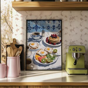 Puede incluir: Una impresi&oacute;n enmarcada de una escena de desayuno con panqueques con bayas, huevos fritos, fruta y caf&eacute;. La obra de arte est&aacute; sobre un mantel blanco con detalles azules y blancos. Una m&aacute;quina de espresso verde est&aacute; a la derecha.