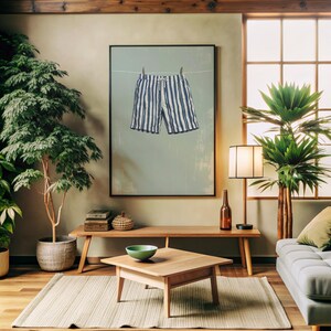 May include: A framed print of blue and white striped shorts hanging on a clothesline. The artwork is displayed in a living room setting with a wooden bench, coffee table, and various plants. The room has a neutral color palette.