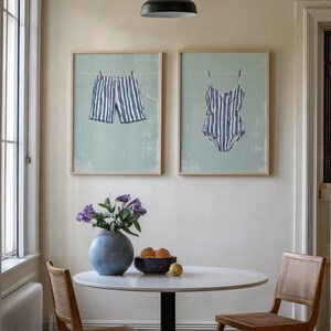 May include: Two framed prints depict blue and white striped swimwear hanging on a clothesline against a light green background. The prints are displayed above a round table with a vase of flowers and a bowl of fruit.