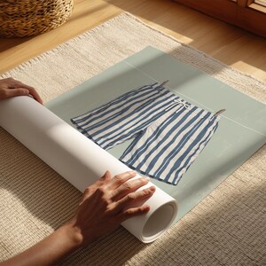 May include: A rolled-up poster featuring a pair of blue and white striped shorts hanging on a clothesline. The poster is being unrolled on a beige rug, with a wicker basket visible in the background.