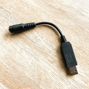 Puede incluir: Un adaptador de cable de alimentación USB a CC negro. El adaptador tiene un conector USB-A y un conector de barril de CC. El cable es negro y está sobre una superficie de madera marrón claro.