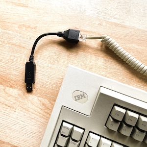 Puede incluir: Un teclado IBM blanco con un cable gris en espiral conectado. El teclado tiene una fila de teclas de función etiquetadas de F1 a F17. El cable está conectado a un adaptador negro con un conector en el extremo.