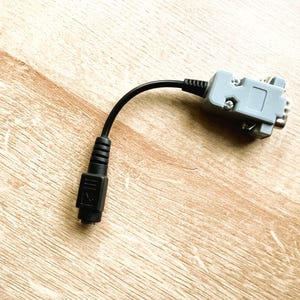 以下が含まれることがあります： 片側に灰色のコネクタ、もう片側に黒いコネクタが付いた黒いケーブル。ケーブルは薄い茶色の木製の表面の上に置かれています。