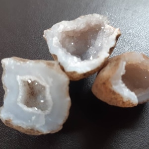 Op de afbeelding: Drie kleine, natuurlijke geodekristallen met een witte buitenkant en een helder, hol interieur. De kristallen zijn bruin aan de buitenkant en hebben een ruwe textuur.