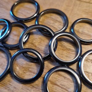 Könnte beinhalten: Eine Ansammlung dunkelgrauer Ringe, möglicherweise Schmuck oder Bastelbedarf, liegt auf einer Holzoberfläche. Die Ringe haben ein glattes, metallisches Aussehen und unterschiedliche Größen. Die Beleuchtung erzeugt Glanzlichter und Schatten.