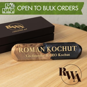 Pode incluir: Uma placa de secretária que combina veios de madeira natural e resina preta. O texto diz ROMAN KOCHUT Co-founder & CEO Kochut. Atrás encontra-se uma caixa retangular preta com o logótipo RWA e uma faixa verde indica OPEN TO BULK ORDERS.