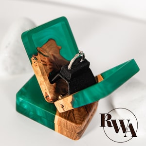 Peut inclure: Un écrin à bagues en bois fait à la main, avec un dessus en résine vert vif et une base en bois naturel. La boîte est ouverte, révélant une bague en argent sur un insert en velours noir. La boîte est conçue pour contenir une bague.