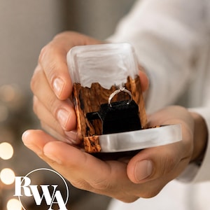 May include: A small rectangular ring box crafted from a combination of natural brown wood and translucent white resin. The interior features a black velvet insert holding a thin silver-toned ring with a small clear stone setting at the top.