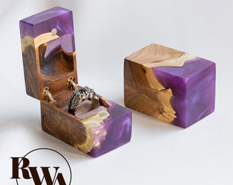 May include: Two rectangular ring boxes crafted from natural wood and translucent purple resin. One box is open, revealing a grey velvet interior holding a textured silver ring with a dark stone. The logo RWA is visible in the bottom left corner.