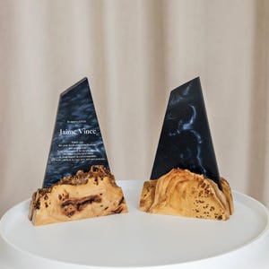 May include: Two custom awards with a dark blue and white marbled design. The awards have a wooden base and a triangular top. One award has text that reads "Jaime Vince". The awards are displayed on a white surface.