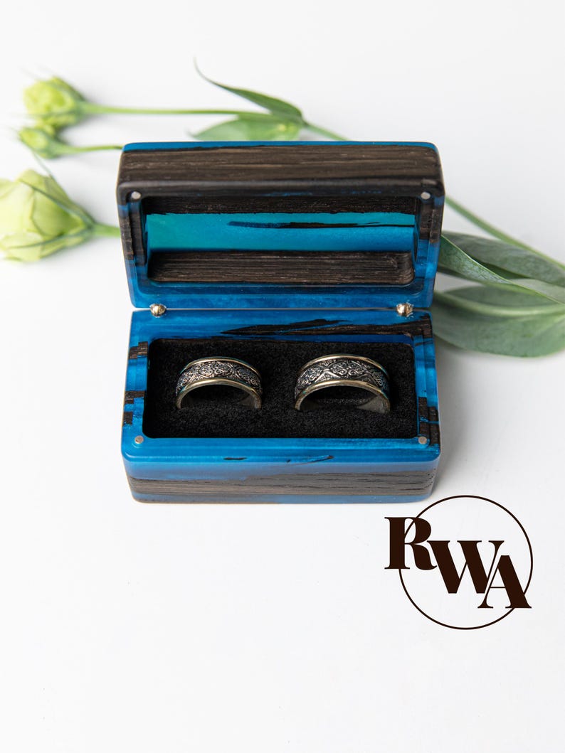 May include: A rectangular ring box crafted from dark wood and vibrant blue resin, featuring a hinged lid and a black interior insert holding two silver-toned metal rings with intricate, embossed floral patterns on their outer surfaces.