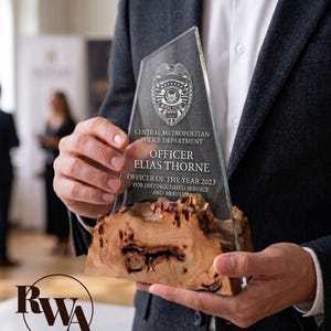 May include: A clear glass award with a wooden base is held by a person. The award features the text "CENTRAL METROPOLITAN POLICE DEPARTMENT OFFICER ELIAS THORNE OFFICER OF THE YEAR 2023 FOR DISTINGUISHED SERVICE AND BRAVERY."