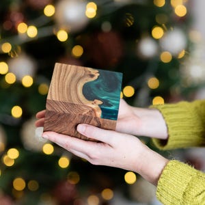 May include: A square wooden box featuring a natural wood grain pattern on one side and a smooth, translucent teal resin inlay on the other. The box has a flat top and sides, showcasing the contrast between the organic wood texture and the resin.