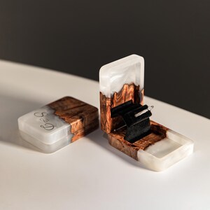 Op de afbeelding: Twee rechthoekige ringdoosjes gemaakt van een combinatie van natuurlijk bruin hout en doorschijnende witte hars. E&eacute;n doosje is geopend en toont een zwarte fluwelen binnenkant met een zilveren ring, terwijl het gesloten doosje de letters S & D bevat.