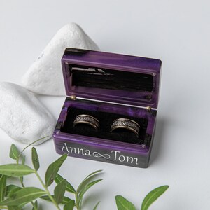 May include: A rectangular purple resin ring box with a hinged lid, featuring the white text Anna and Tom with an infinity symbol. The interior is lined with black velvet and holds two silver-toned metal rings with intricate engraved patterns.