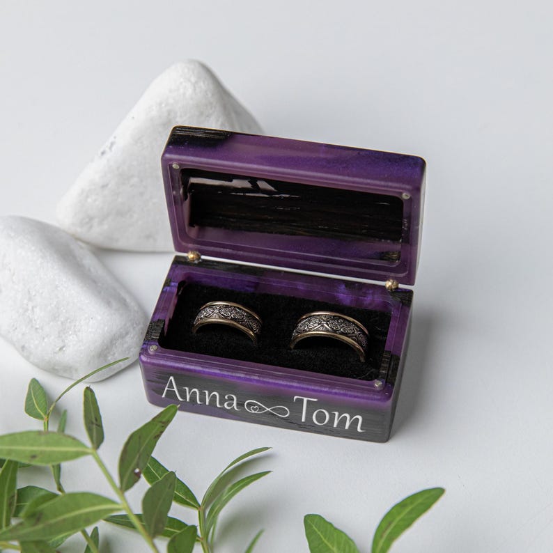 May include: A rectangular purple resin ring box with a hinged lid, featuring the white text Anna and Tom with an infinity symbol. The interior is lined with black velvet and holds two silver-toned metal rings with intricate engraved patterns.