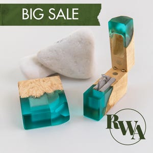Può includere: Un portaanelli in legno chiaro e resina turchese traslucida, con coperchio incernierato e interno in velluto grigio contenente un anello d'argento. Il testo BIG SALE appare su un banner verde e le lettere RWA sono racchiuse in un cerchio.
