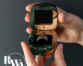 May include: A small, square ring box crafted from dark green resin and natural wood burl. The box is open, revealing a black velvet interior holding a silver-toned ring with a textured band. The logo RWA is visible in the bottom left corner.